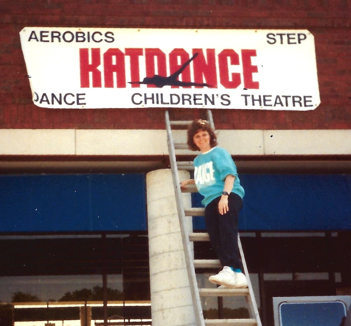 Photo of Katie on a ladder next to the Katdance Children's Theatre sign.
